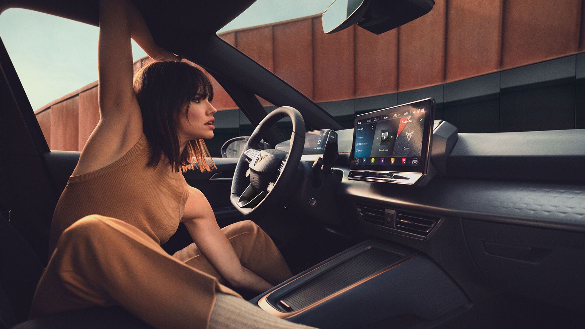 Oseba sedi v udobni elektronsko opremljeni notranjosti avtomobila in z roko drži odprto strešno okno. Notranjost avtomobila je moderna, z velikim zaslonom na armaturni plošči. Zunaj je vidna stavba s kovinsko fasado.