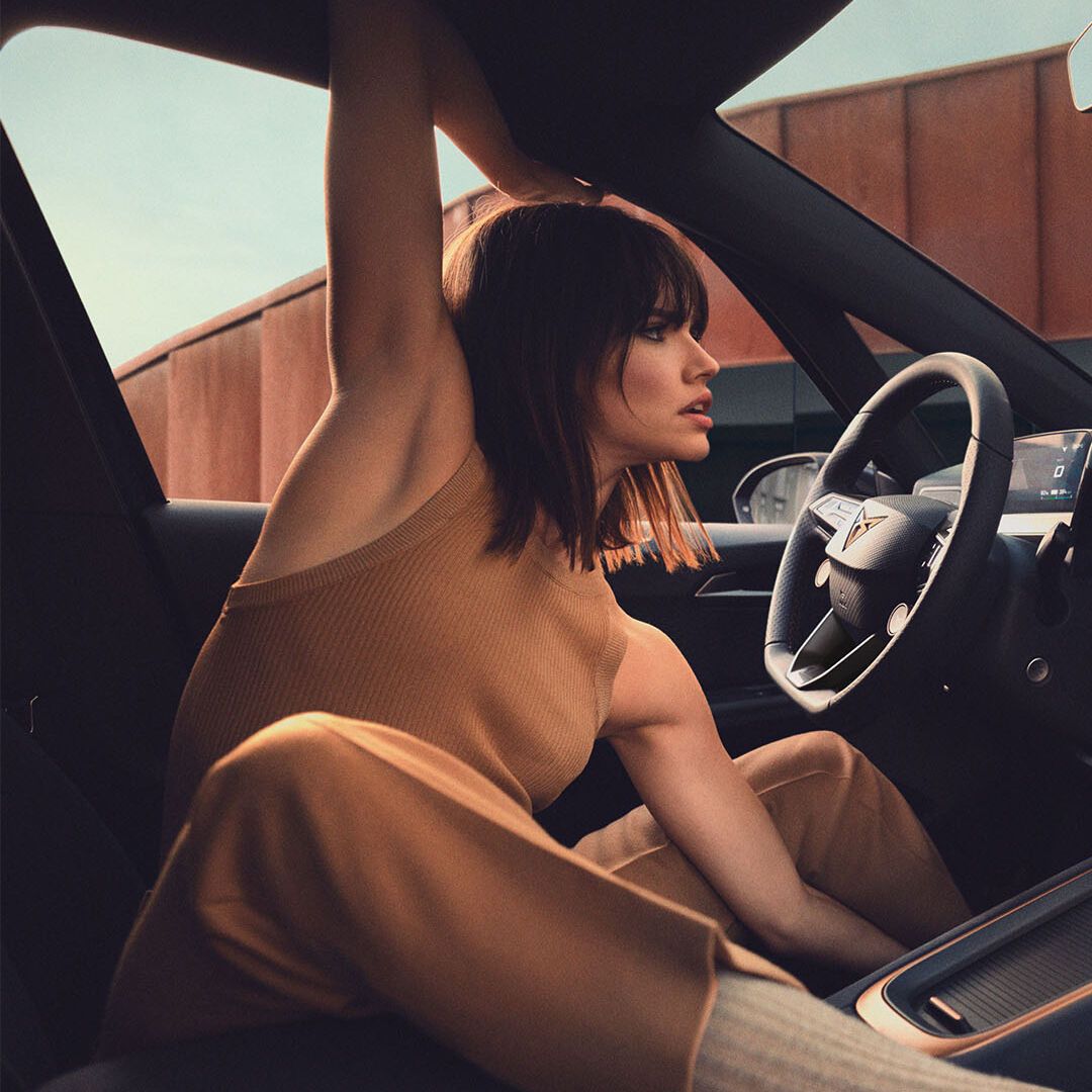 Oseba sedi v udobni elektronsko opremljeni notranjosti avtomobila in z roko drži odprto strešno okno. Notranjost avtomobila je moderna, z velikim zaslonom na armaturni plošči. Zunaj je vidna stavba s kovinsko fasado.