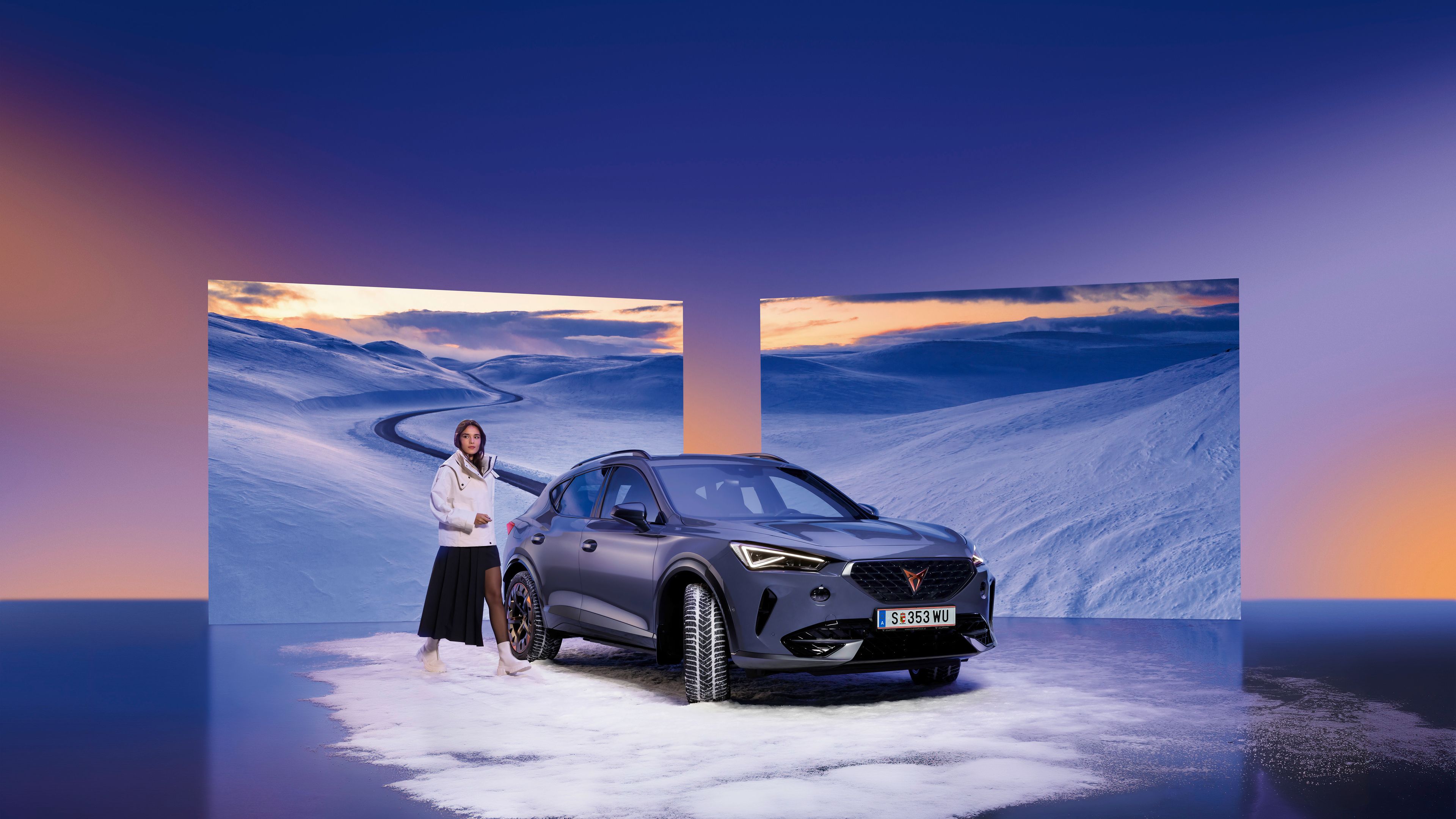 Temno siv CUPRA Formentor stoji na zasneženi površini z dvema velikima ekranoma v ozadju, ki prikazujeta zasneženo pokrajino. Poleg avtomobila je ženska v belem oblačilu. Ozadje je modro-vijolično.