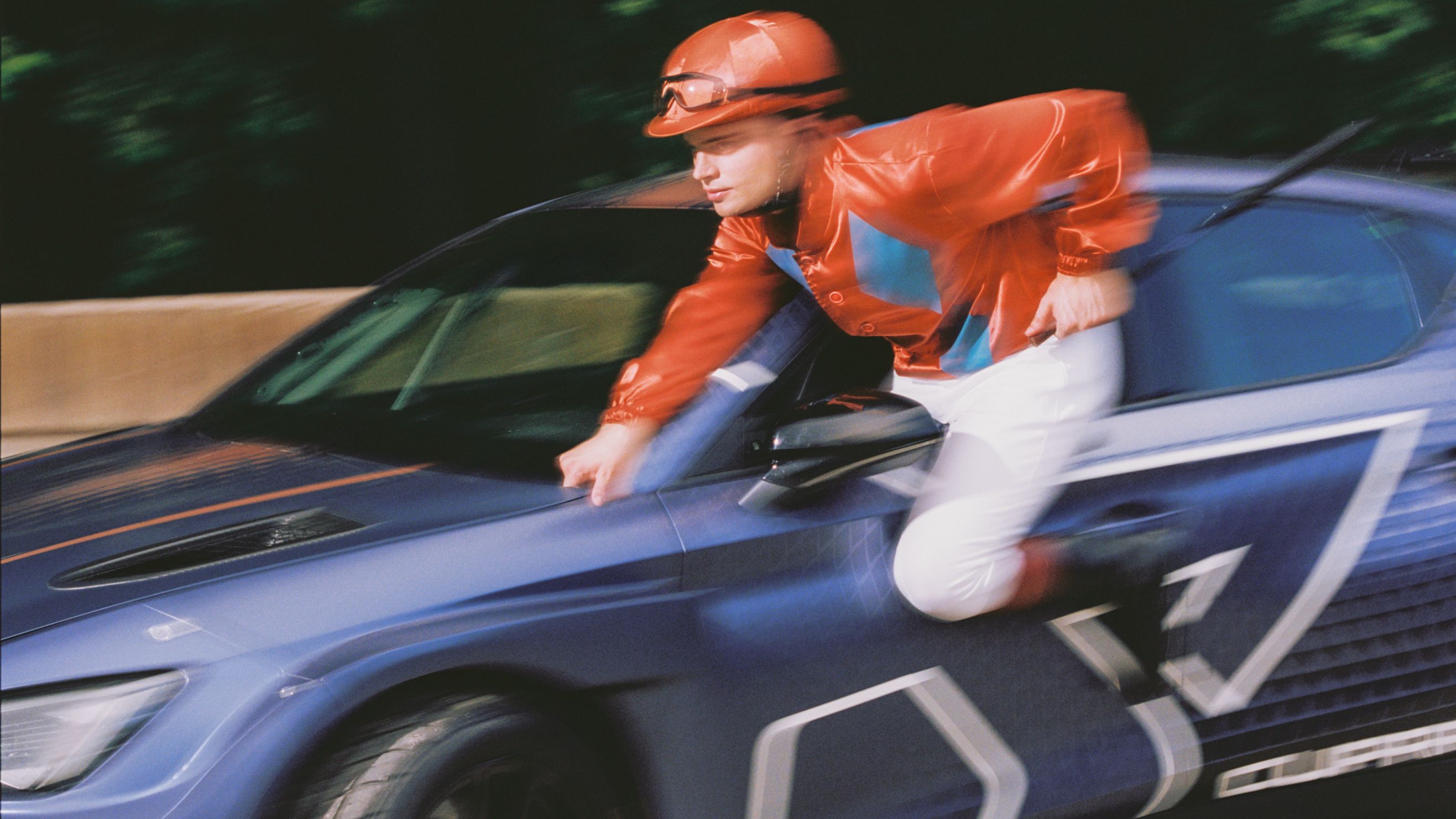 Oseba v zaščitni čeladi in oranžni jakni se v gibanju naslanja na modro športno vozilo znamke CUPRA. Dinamičen posnetek z zamegljenim ozadjem poudarja hitrost in energijo prizora.