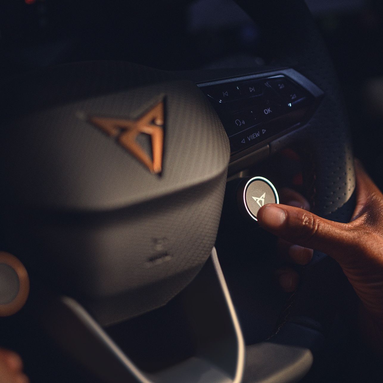 Oseba pritiska na osvetljen gumb z ikono volana na volanu avtomobila Cupra Born. Volan je temen z osvetljenimi kontrolami. Osvetlitev je šibka, kar ustvarja prijetno in intimno vzdušje.