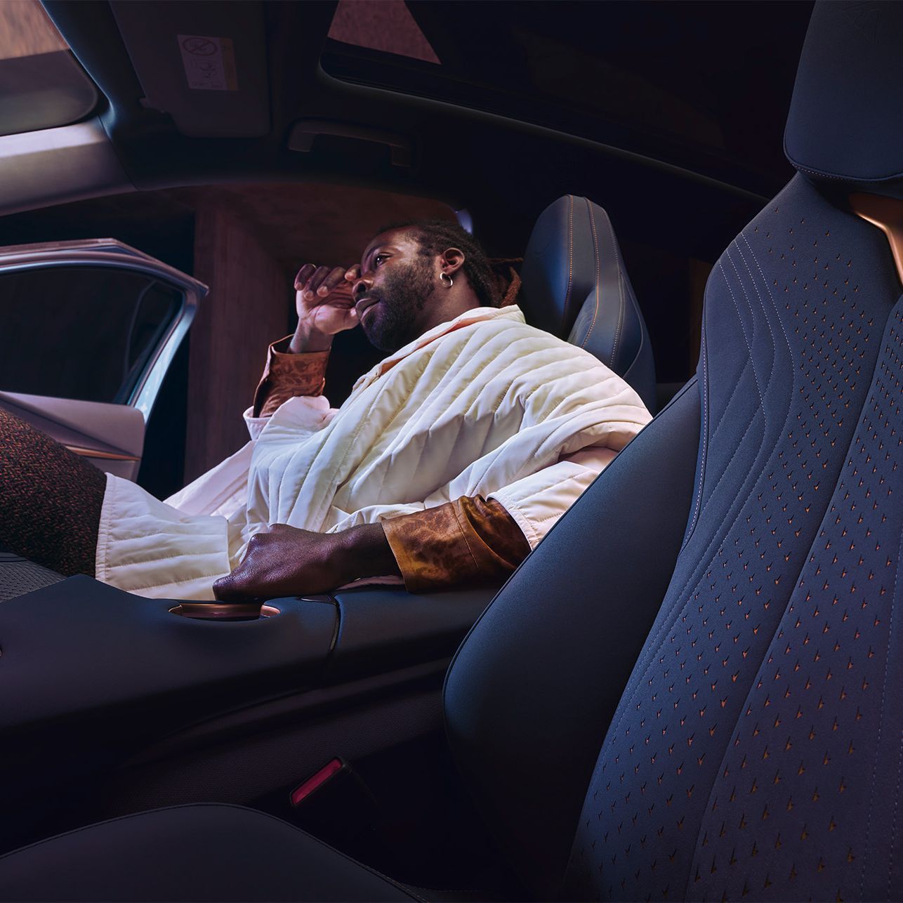 Moški sedi v Cupra Tavascan avtomobilu, z odprtimi vrati na sovoznikovi strani. Na sedežu je oblečena v belo jakno in zdi se, da se sprošča. Avto ima sodobno oblikovan notranji prostor.