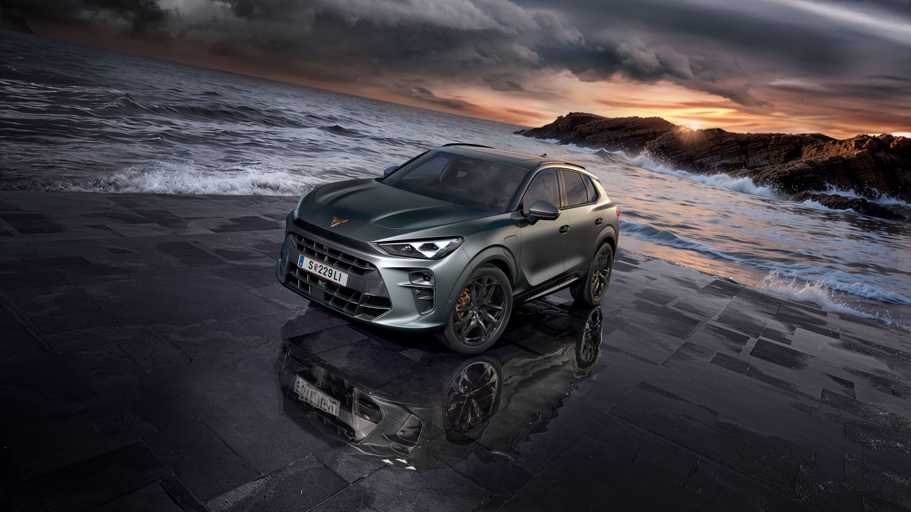 Sodobna temno sivo vozilo CUPRA Terramar, s črnimi platišči, na mokrem tlakovcu ob obali z oblačnim nebom v ozadju. Morje, valovi in skalnata obala se raztezajo v ozadju.