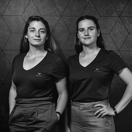 Dve igralki padel tenisa, oblečeni v črne majice. Ozadje je geometrijsko s temnimi odtenki.