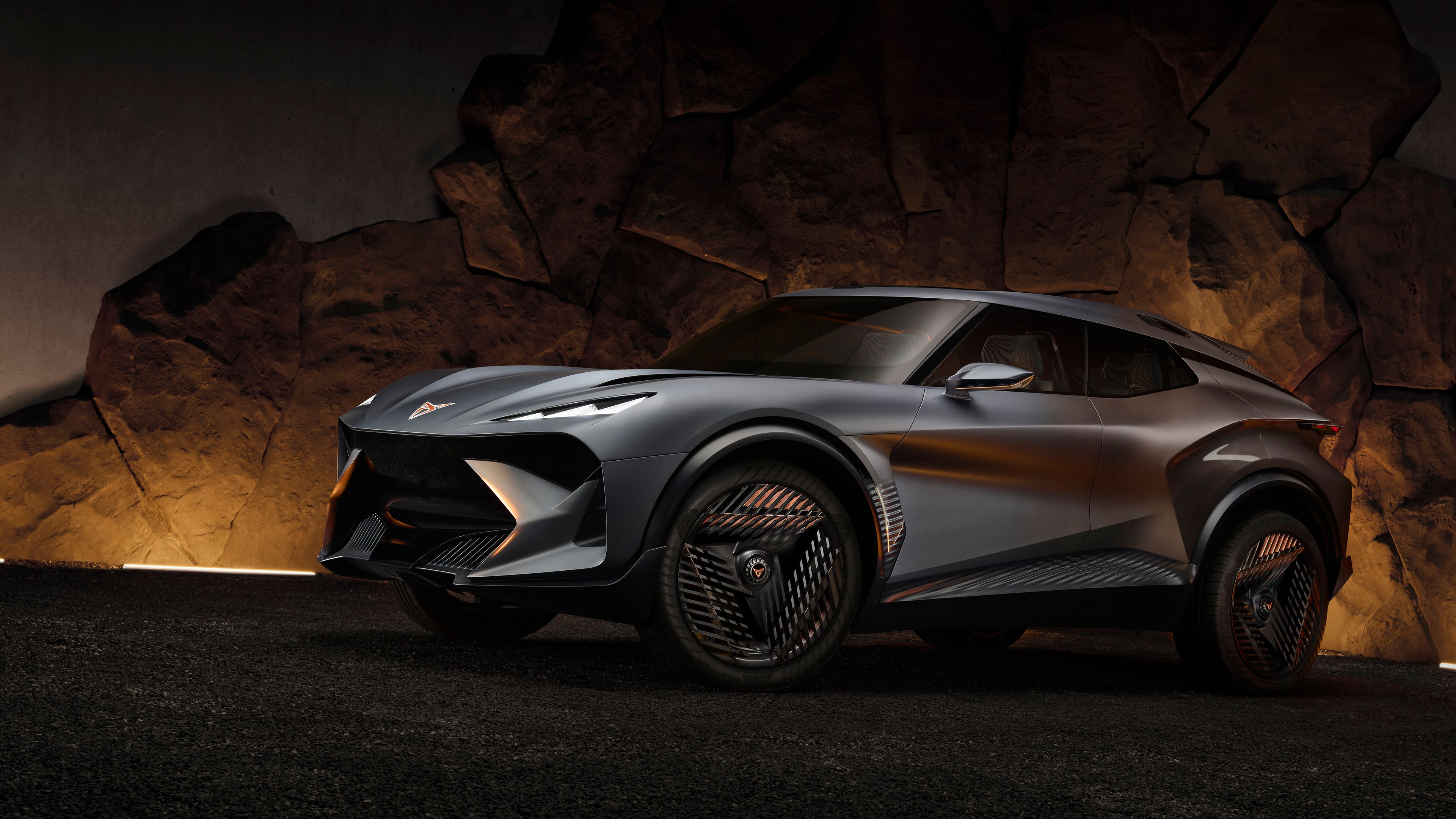 Nov siv športni avtomobil CUPRA Tindaya je parkirana pred kamnito steno. Avto ima velikanske platišča z edinstveno zasnovo in aerodinamične linije. Osvetljava poudarja moderno oblikovanje vozila.