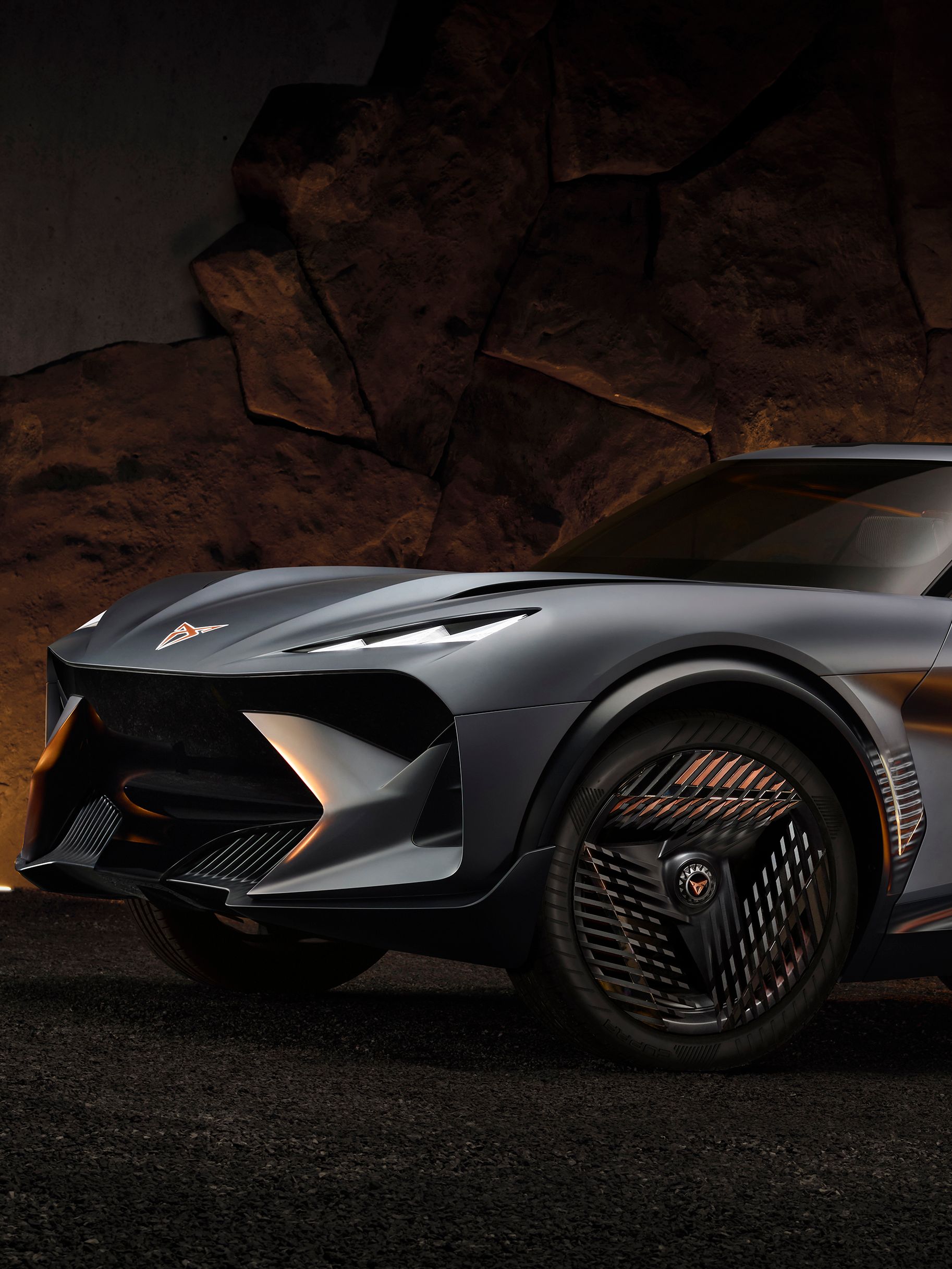 Nov siv športni avtomobil CUPRA Tindaya je parkirana pred kamnito steno. Avto ima velikanske platišča z edinstveno zasnovo in aerodinamične linije. Osvetljava poudarja moderno oblikovanje vozila.