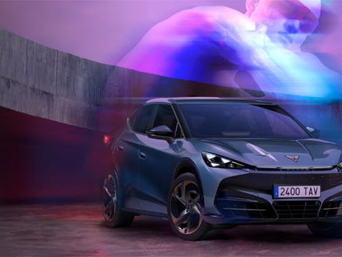 Slika prikazuje temno moder Cupra Tavascam postavljen pred moderni, ukrivljen zid z vijoličnimi in modrimi svetlobnimi efekti v ozadju. Na desni strani slike je abstraktni logotip Cupra v beli barvi. Ozadje je zamegljeno z odtenki vijolične, rožnate in sive barve.