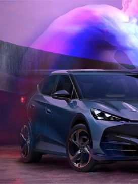 Slika prikazuje temno moder Cupra Tavascam postavljen pred moderni, ukrivljen zid z vijoličnimi in modrimi svetlobnimi efekti v ozadju. Na desni strani slike je abstraktni logotip Cupra v beli barvi. Ozadje je zamegljeno z odtenki vijolične, rožnate in sive barve.