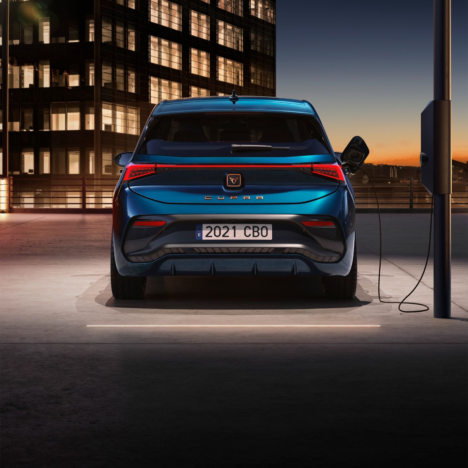 Električni avto CUPRA Born modre barve se polni na polnilni postaji na zunanjem parkirišču. V ozadju je osvetljena moderna stavba.
