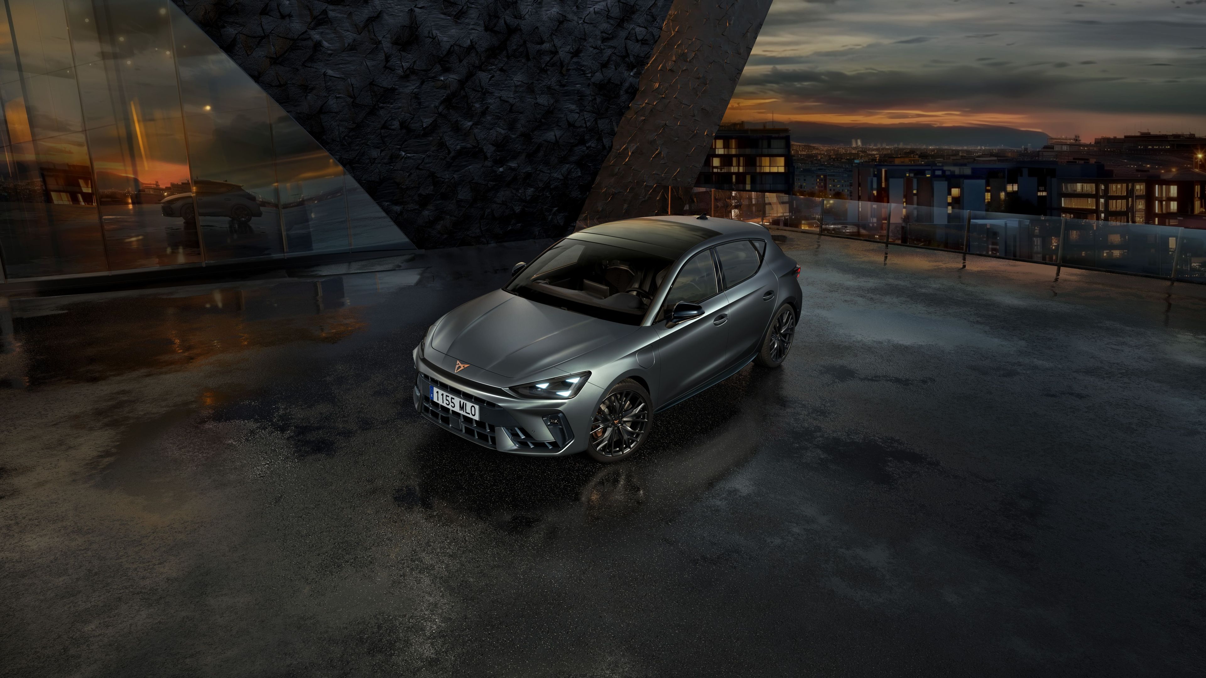 Sivi športni avto CUPRA je parkiran na mokrem parkirišču ob moderni stavbi. Nebo je oblačno, v ozadju pa je videti mesto ob sončnem zahodu.