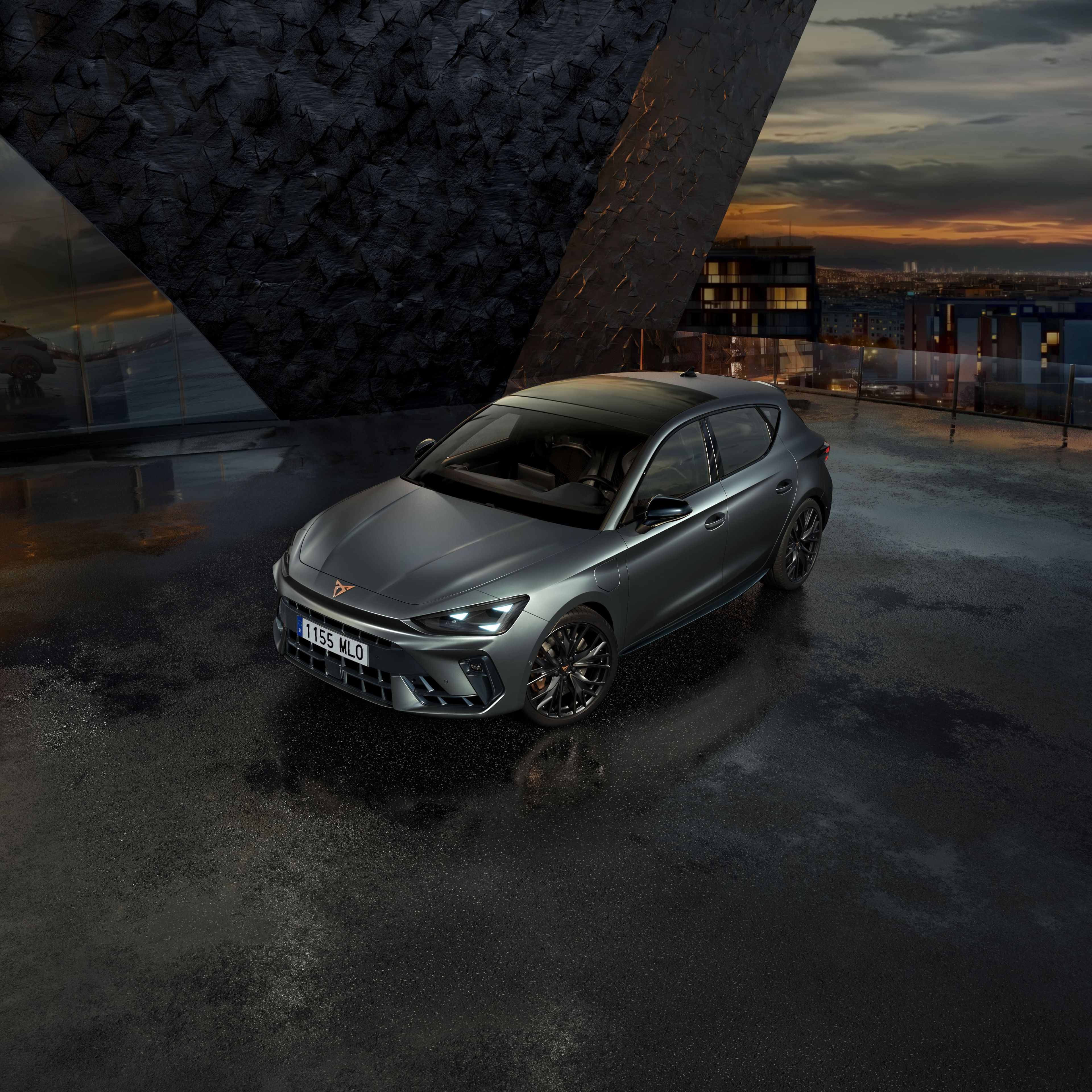 Sivi športni avto CUPRA je parkiran na mokrem parkirišču ob moderni stavbi. Nebo je oblačno, v ozadju pa je videti mesto ob sončnem zahodu.