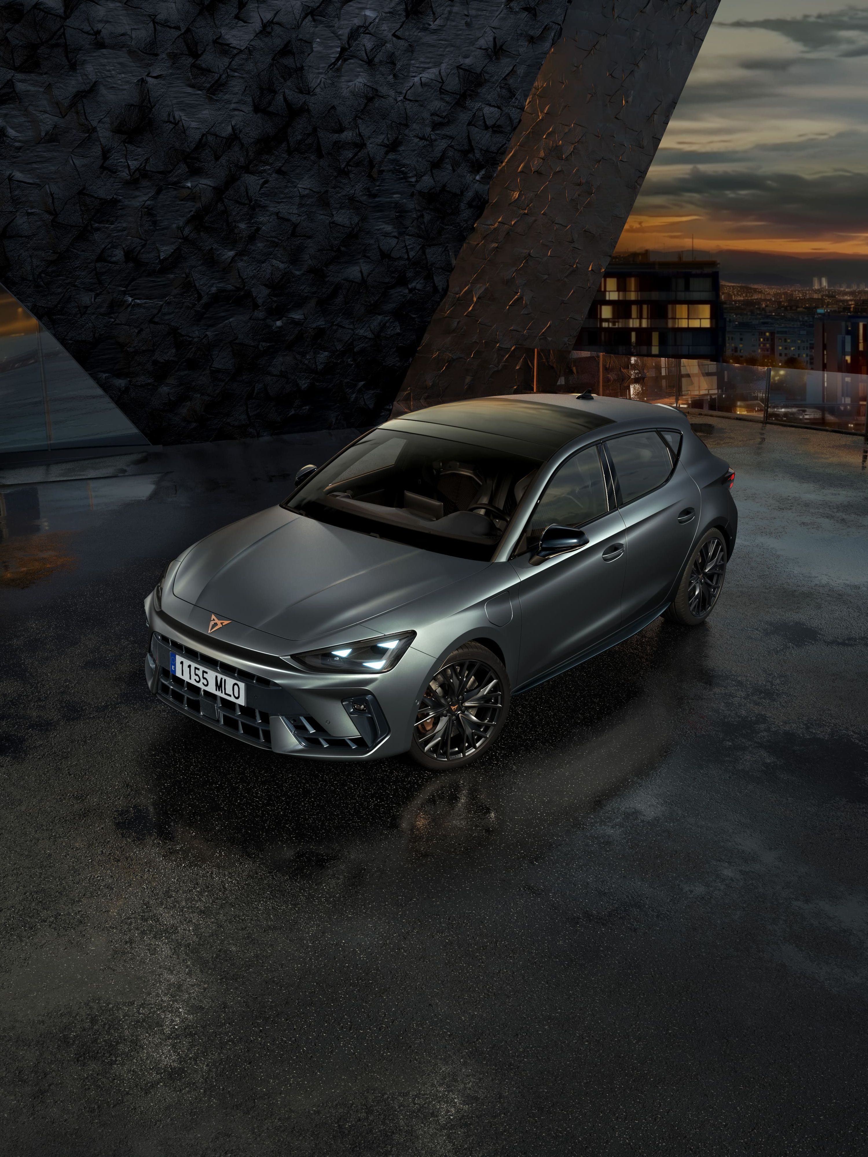 Sivi športni avto CUPRA je parkiran na mokrem parkirišču ob moderni stavbi. Nebo je oblačno, v ozadju pa je videti mesto ob sončnem zahodu.