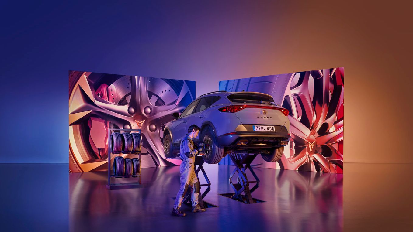 CUPRA Formentor je dvignjen na dvigalkah v studiu z umetniško osvetljenim ozadjem. V ozadju sta dva velika zaslona z abstraktnimi podobami. Oseba dela na avtomobilu z orodjem.