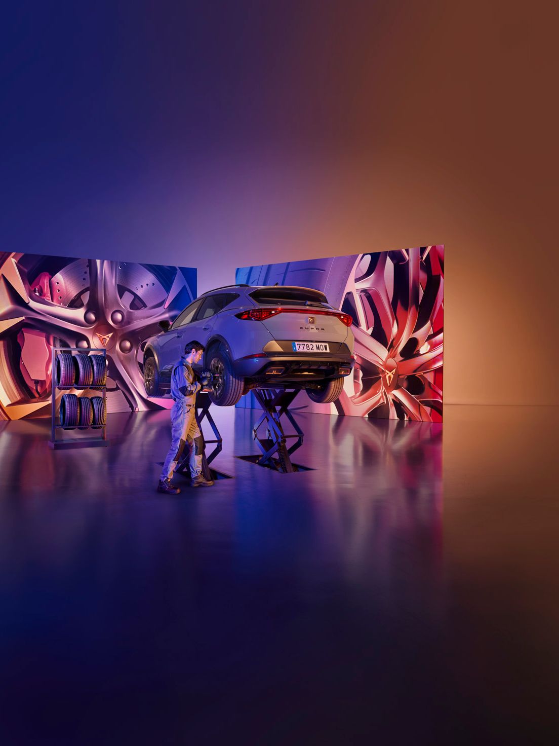 CUPRA Formentor je dvignjen na dvigalkah v studiu z umetniško osvetljenim ozadjem. V ozadju sta dva velika zaslona z abstraktnimi podobami. Oseba dela na avtomobilu z orodjem.
