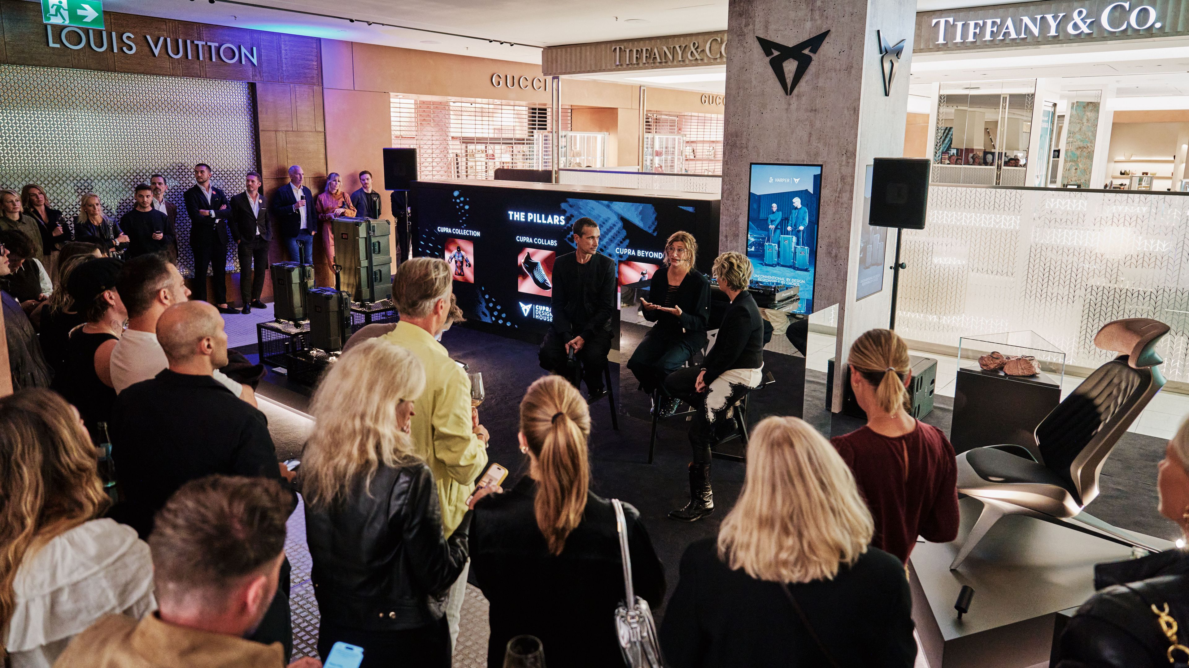 Prizor prikazuje skupino ljudi na dogodku v nakupovalnem centru. Nekateri udeleženci sedijo, medtem ko drugi stojijo in opazujejo predstavitev na velikem zaslonu.