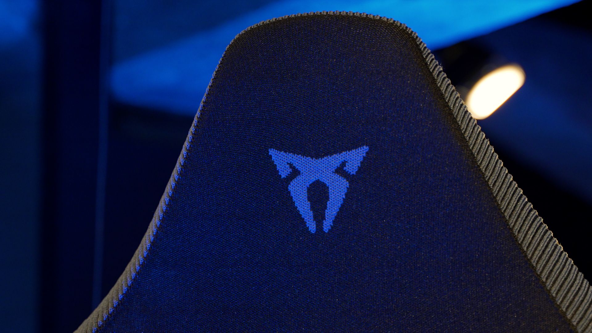 Podrobna fotografija zgornjega dela hrbtnega naslona avtomobilskega sedeža z modro osvetlitvijo, na katerem je videti logotip znamke CUPRA. Stol ima črno oblazinjenje.