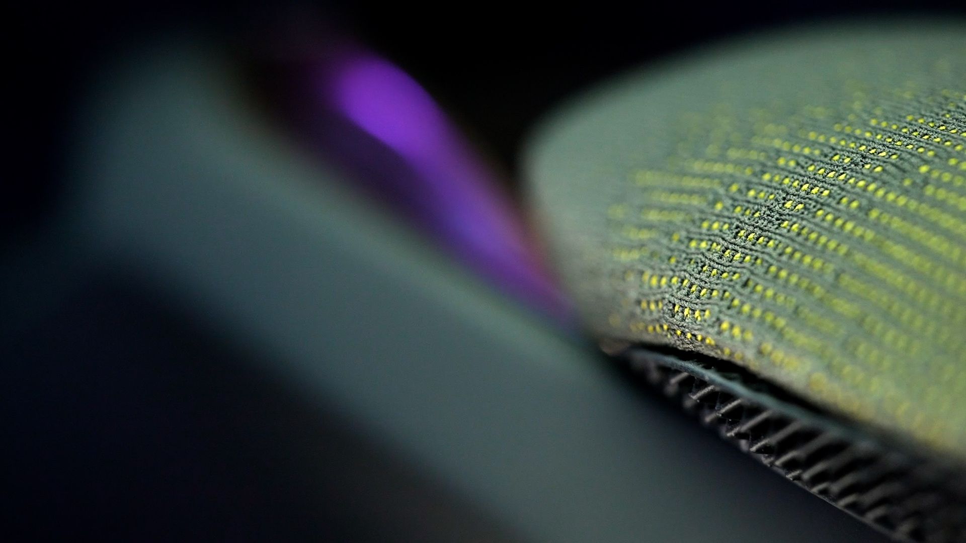 Zelo podroben posnetek tekstilne površine v zeleni barvi. V ozadju je zamegljen vijoličen odsev. Tekstilna struktura je izrazita in vidne so posamezne niti.