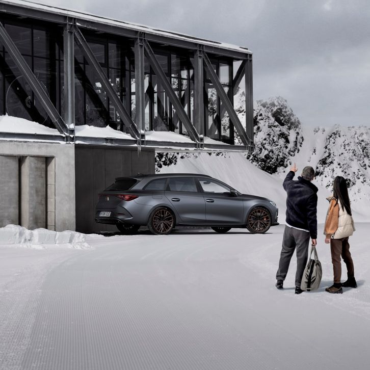 Na sliki sta moški in ženska, ki stojita na sneženi površini blizu sodobne zgradbe s steklenimi in kovinskimi elementi. CUPRA Leon PA je parkiran ob stavbi.