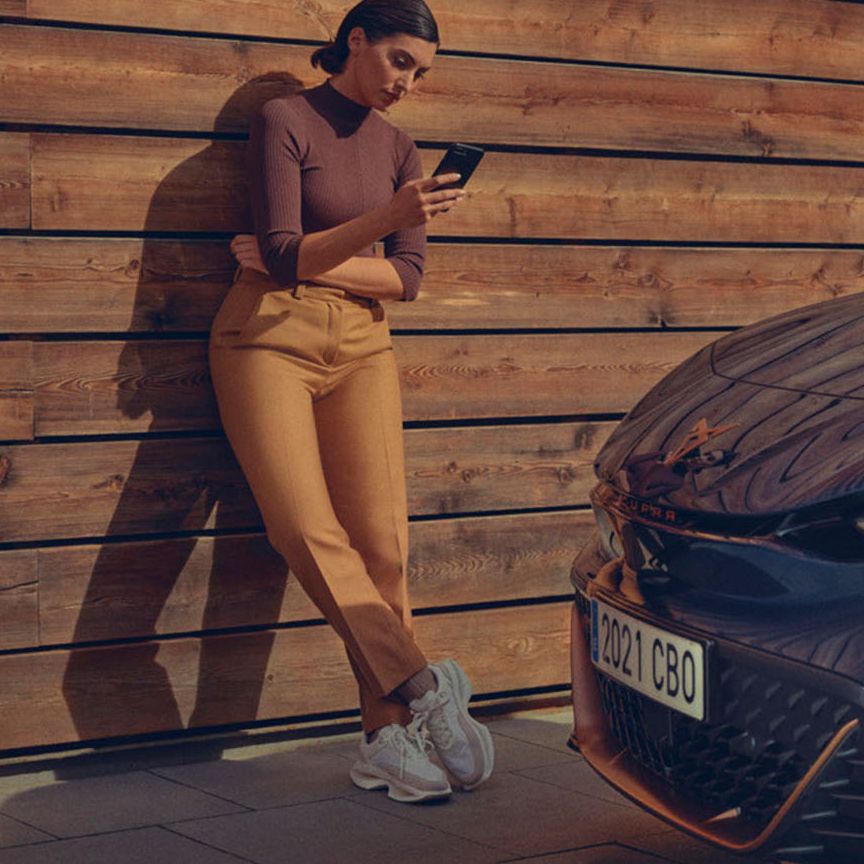 Ženska stoji naslonjena na leseno steno in gleda v svoj mobilni telefon. Ob njej je parkiran moder CUPRA Born