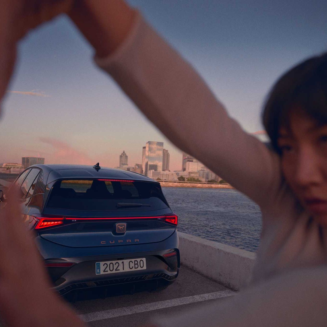 Oseba z zamegljenim obrazom je iztegnila roke v obliki okvirja pred avtomobilom CUPRA Born, parkiranim ob reki. Avtomobil je temno modre barve. V ozadju se vidijo mestne stavbe in obarvano nebo ob sončnem zahodu.