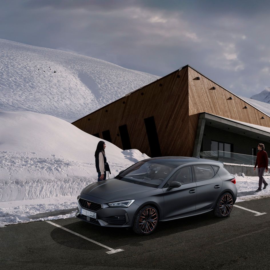 CUPRA Leon 5D je parkiran na asfaltiranem parkirišču pred sodobno zgradbo, ki stoji v zasneženi pokrajini. V bližini avtomobila sta dve osebi – ženska stoji, moški pa hodi proti avtomobilu. V ozadju so zasnežene gore in oblačno nebo.