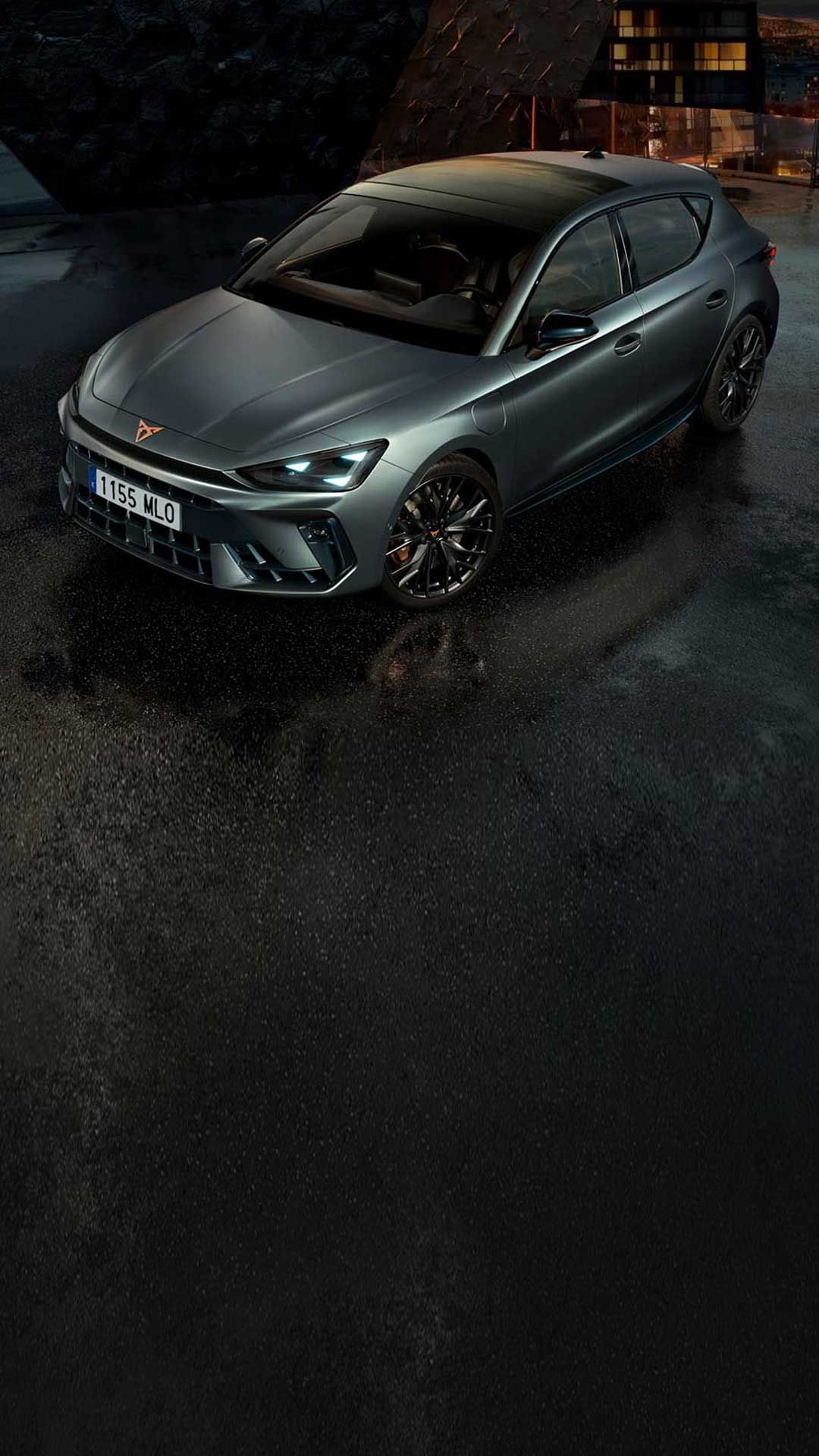CUPRA Leon v kovinsko sivi barvi stoji na mokri strešni terasi ob mraku, v ozadju se blešči razsvetljeno mesto. 