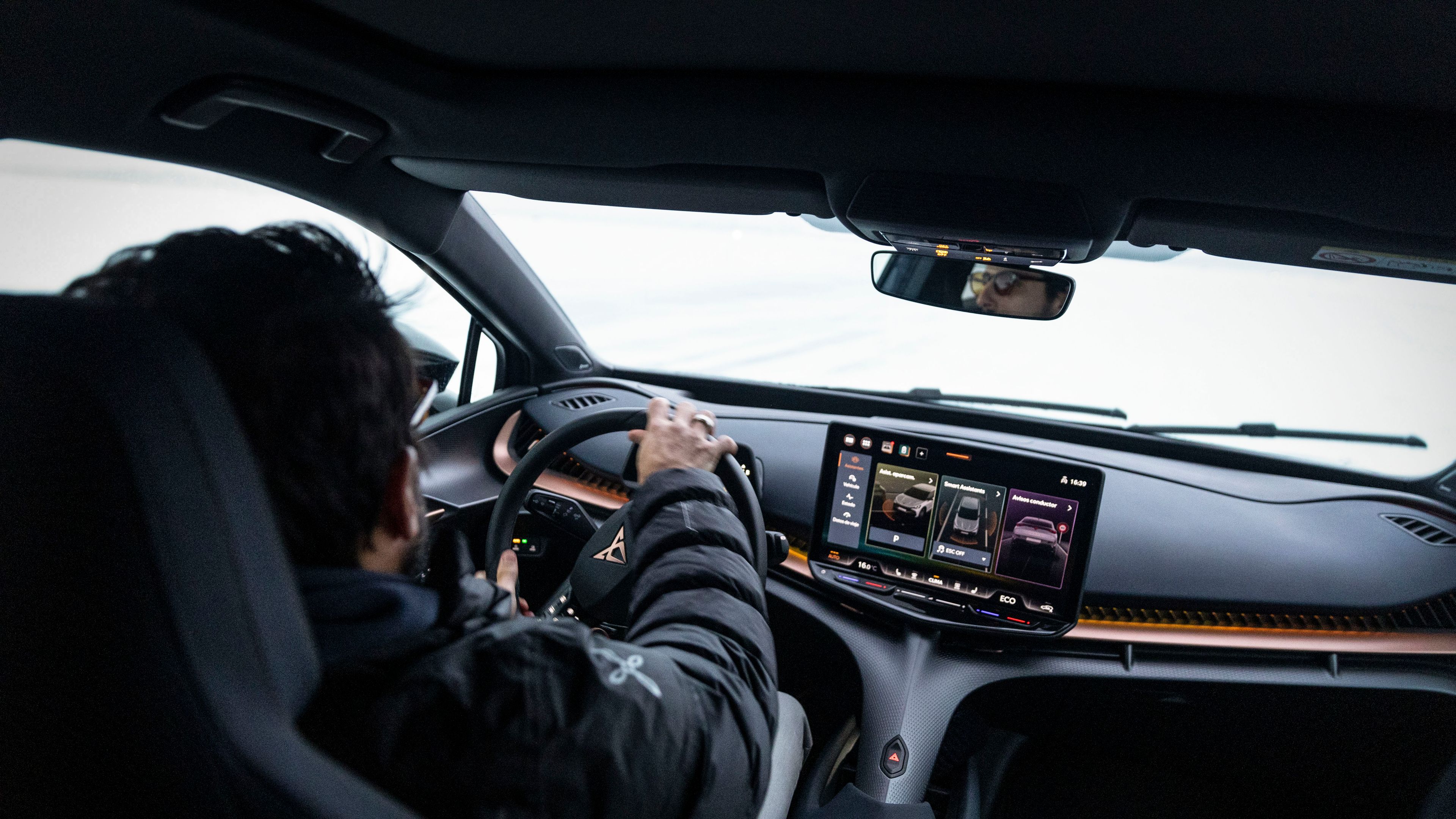 Lucas di Grassi vozi avtomobil CUPRA Tavascan, medtem ko gleda na velik osrednji zaslon na armaturni plošči. Notranjost avtomobila je moderna in športna, z digitalnimi prikazovalniki in elegantnimi sedeži.