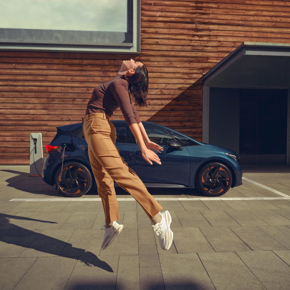 Ženska v rjavih hlačah in temni majici skače v zrak pred modrim avtomobilom CUPRA Born parkiranim na dovozu. Ozadje je lesena stavba.
