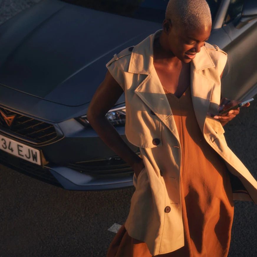 Ženska v bež plašču stoji na soncu pred parkiranim mat sivim vozilom CUPRA Leon 5D in gleda v telefon. 