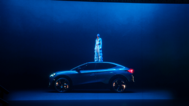 Na fotografiji oseba stoji na strehi modrega avtomobila znamke CUPRA v temnem, osvetljenem okolju. Avtomobil ima sodoben dizajn. 