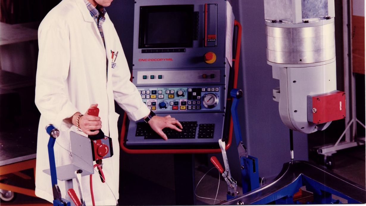 Oseba v laboratorijskem plašču stoji pred računalniško krmiljenim strojem in upravlja s kontrolami. Na zaslonu stroja so prikazani različni gumbi in tipkovnica. Slika je stara približno 50 let.