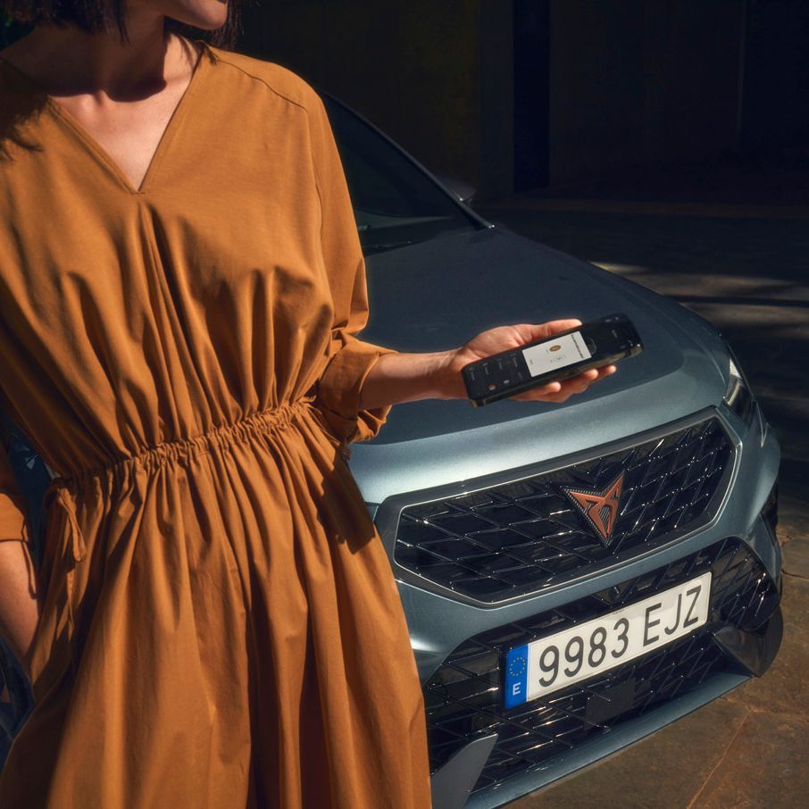 Ženska v oranžni obleki stoji pred sivim avtomobilom CUPRA Ateca i drži pametni telefon. 