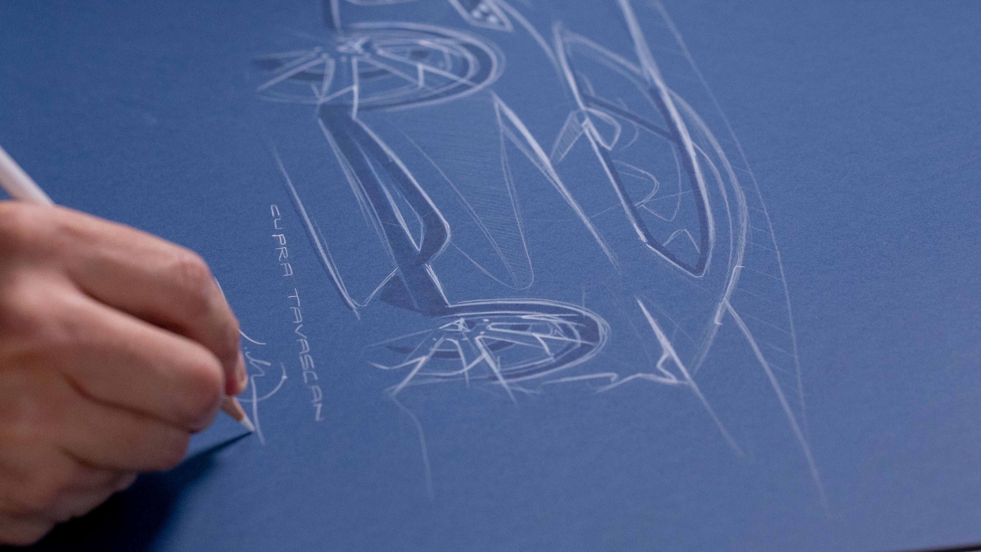 Risba avtomobila na modrem papirju. Načrt vključuje profil vozila CUPRA Tavascan s podrobnostmi kot so kolesa in druge oblikovne značilnosti. Oseba na levi strani drži svinčnik in riše skico.