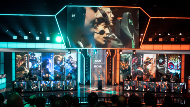 Prostor z občinstvom, ki gleda na oder, kjer je več računalniških postaj z igralci video iger. Zasloni prikazujejo like in statistike iz video igre. Osvetljenost je močna, z modro in oranžno tematiko.