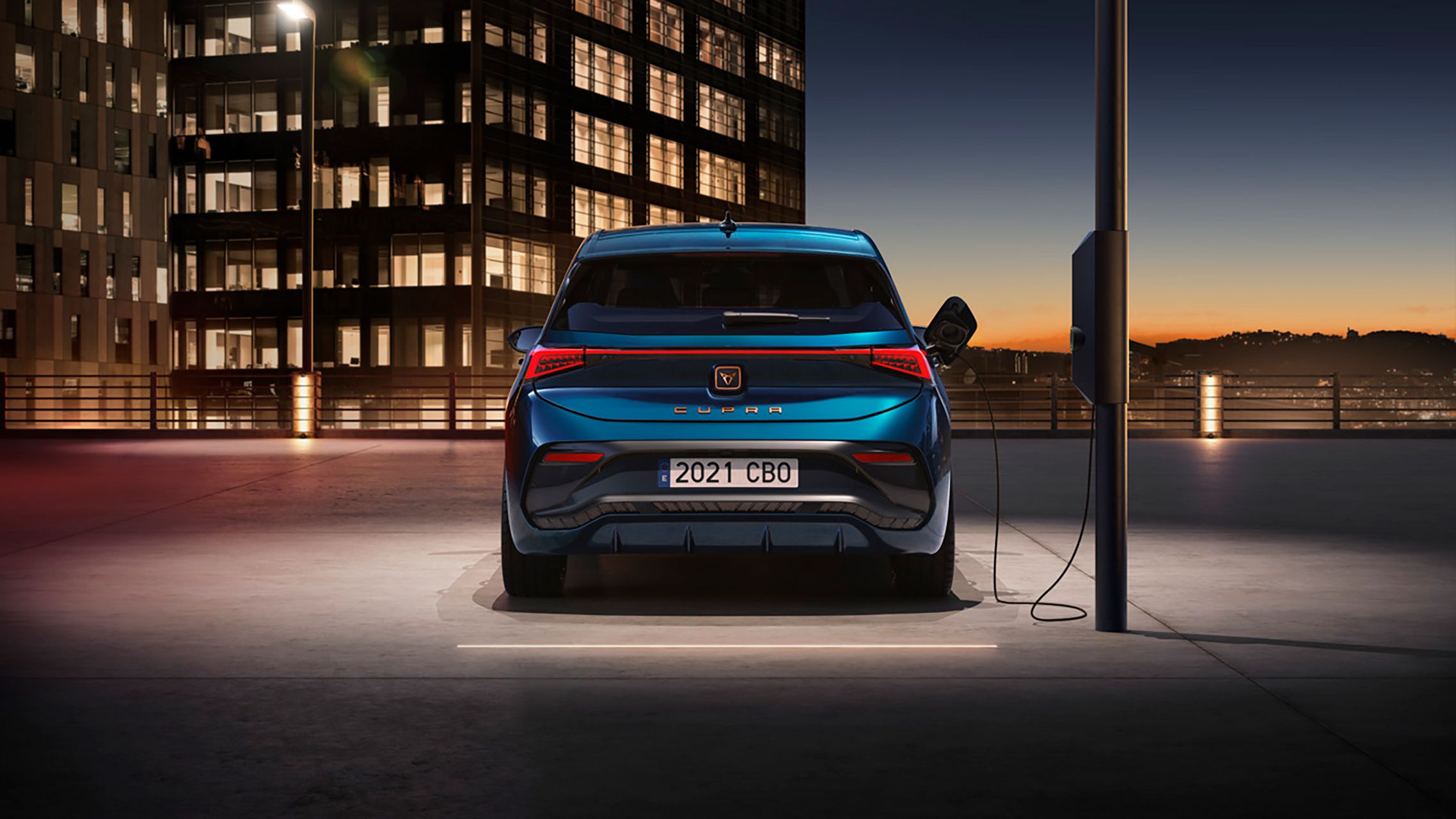 Električni CUPRA Born, modre barve, parkiran v mestnem okolju ob električni polnilni postaji med sončnim zahodom. V ozadju so visoke stavbe. 