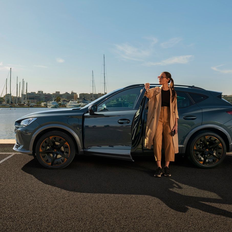 Oseba stoji ob odprtih vratih sivega vozila CUPRA Formentor, parkiranega ob vodi s pristaniščem v ozadju. 