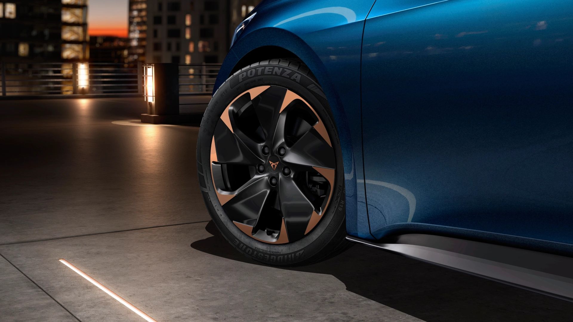 Delni posnetek CUPRA Borna na parkirišču v mestnem okolju. Poudarek je na sprednji levi pnevmatiki, ki ima edinstvena platišča v črni in zlati barvi.