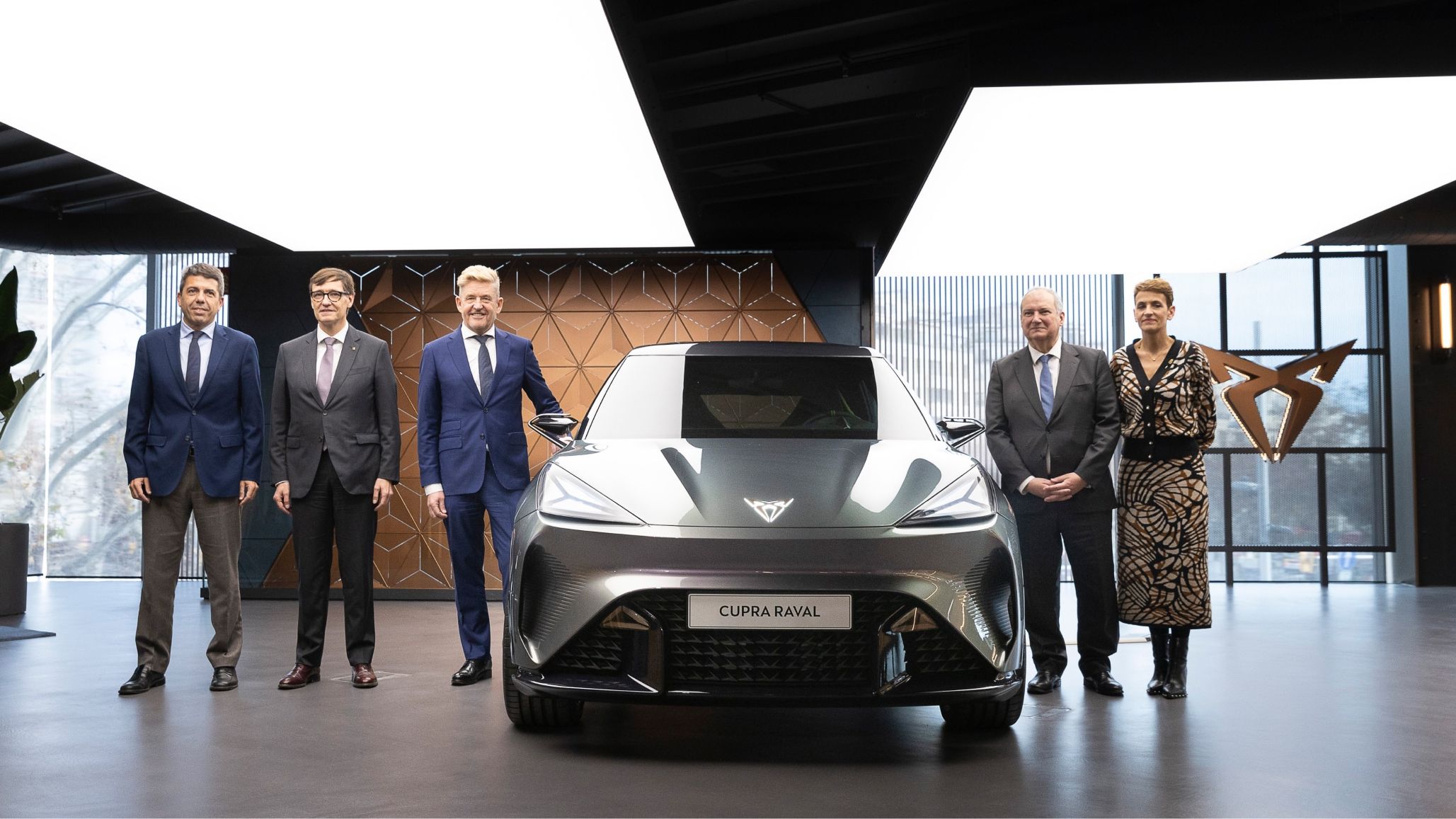 Več ljudi stoji ob izpostavljenem avtomobilu CUPRA Raval. Notranjost je sodobna z geometrijskimi vzorci in velikimi steklenimi površinami. Avto je temno sive barve.