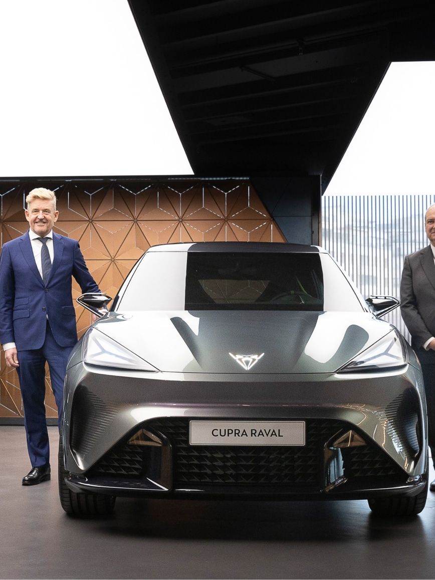 Več ljudi stoji ob izpostavljenem avtomobilu CUPRA Raval. Notranjost je sodobna z geometrijskimi vzorci in velikimi steklenimi površinami. Avto je temno sive barve.