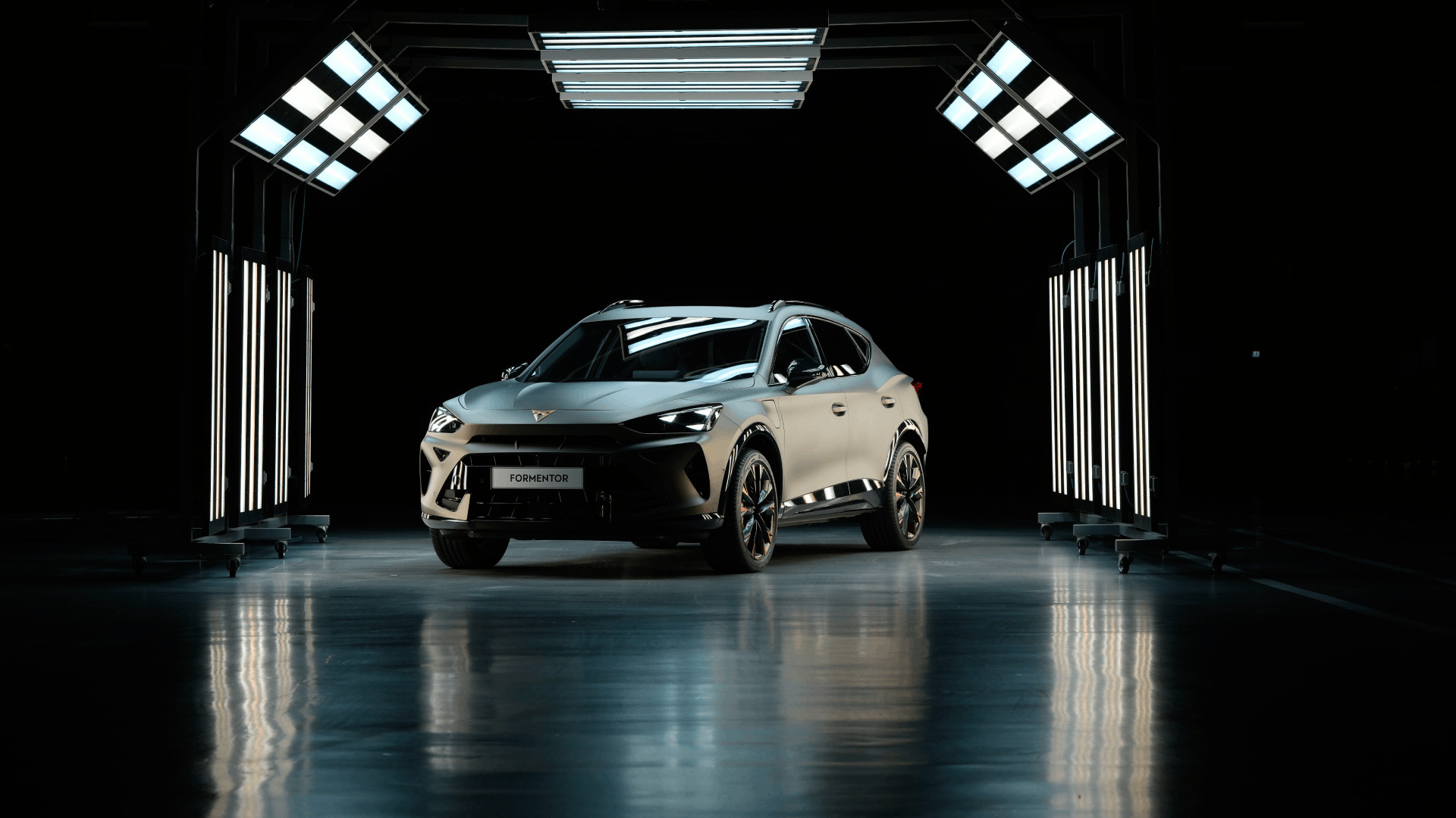Sodoben športni avtomobil CUPRA Formentor je osvetljen v temnem prostoru, obdanim s svetilkami. Avtomobil ima aerodinamičen dizajn in športne značilnosti. Pritlične luči ustvarjajo sijaj na mokri tleh.
