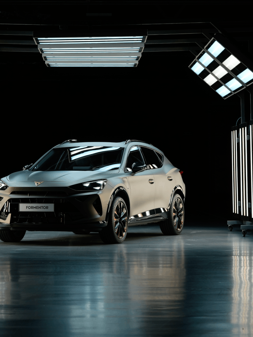 Sodoben športni avtomobil CUPRA Formentor je osvetljen v temnem prostoru, obdanim s svetilkami. Avtomobil ima aerodinamičen dizajn in športne značilnosti. Pritlične luči ustvarjajo sijaj na mokri tleh.