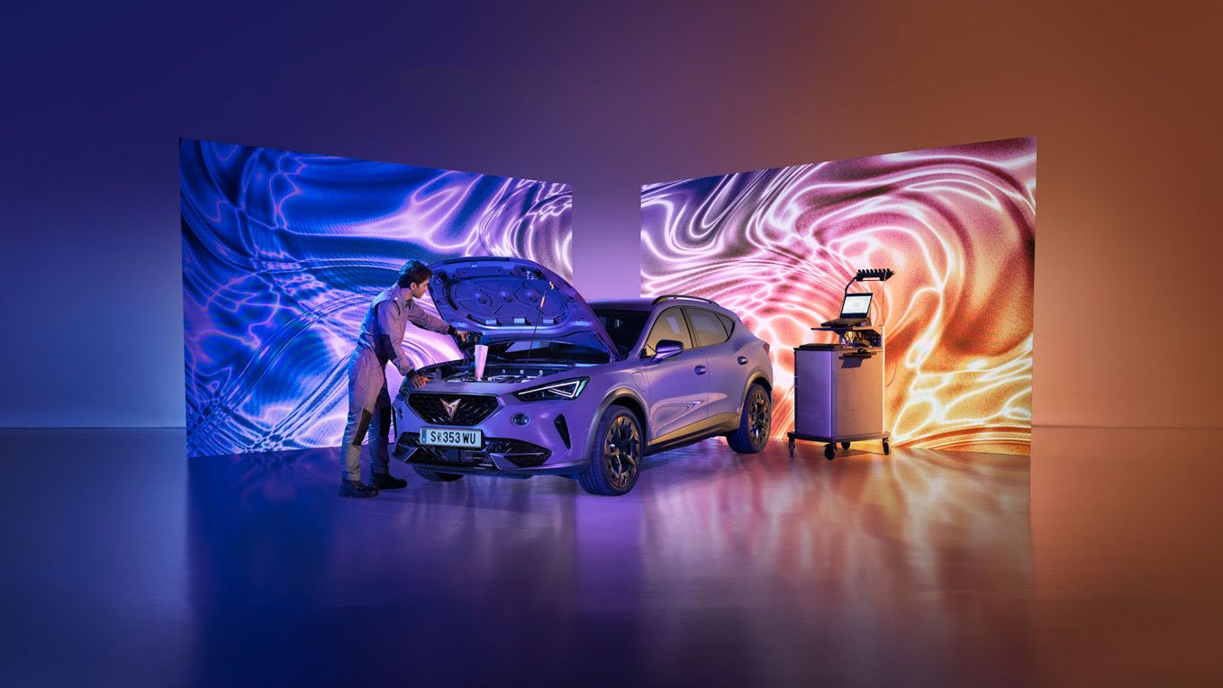 Moški pregleduje motor avtomobila, parkiranega v svetlem studiu s psihodeličnimi vzorci na zaslonih. Poleg vozila je voziček z orodjem, vzdušje pa je moderno in tehnološko napredno.