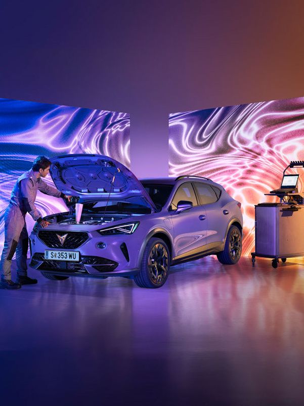 Moški pregleduje motor avtomobila, parkiranega v svetlem studiu s psihodeličnimi vzorci na zaslonih. Poleg vozila je voziček z orodjem, vzdušje pa je moderno in tehnološko napredno.