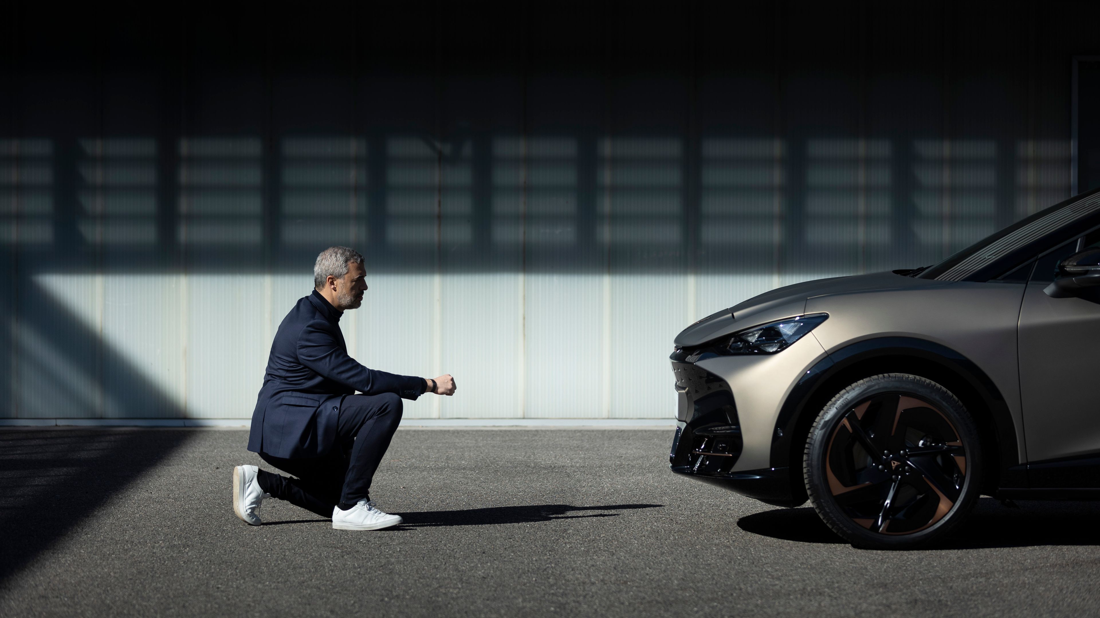 Oblikovalec Jorge Díez v temni obleki kleči ob parkiranem avtomobilu CUPRA Tavascan na ulici. Avto je bronaste barve in sodobnega videza. 