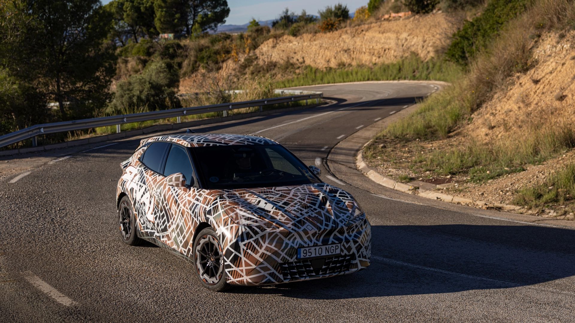 Zamaskiran prototip avtomobila CUPRA Raval Dynamic vozi po vijugasti podeželski cesti ob sončnem dnevu. Pokrajina je kamnita in polna zelenja. 