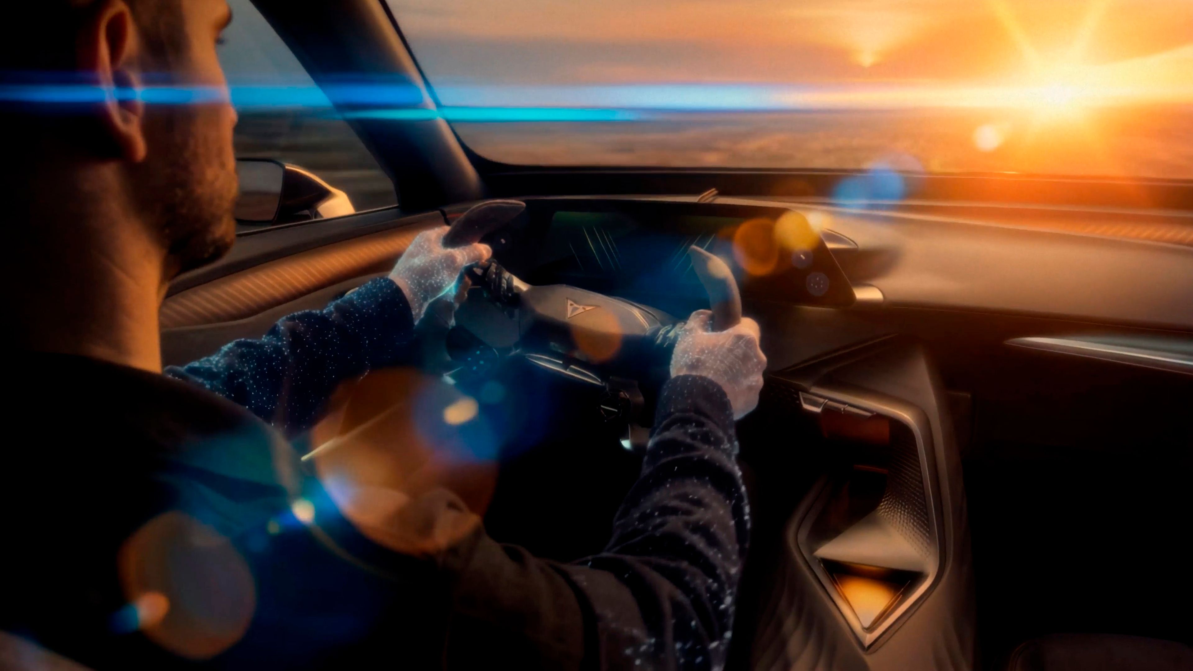Voznik v vozilu Tindaya, ki z rokami drži volan. Spredaj je vidno sonce, žarki sijejo v vozilo. Pogled iz zadnjega dela avta na voznika in armaturno ploščo.