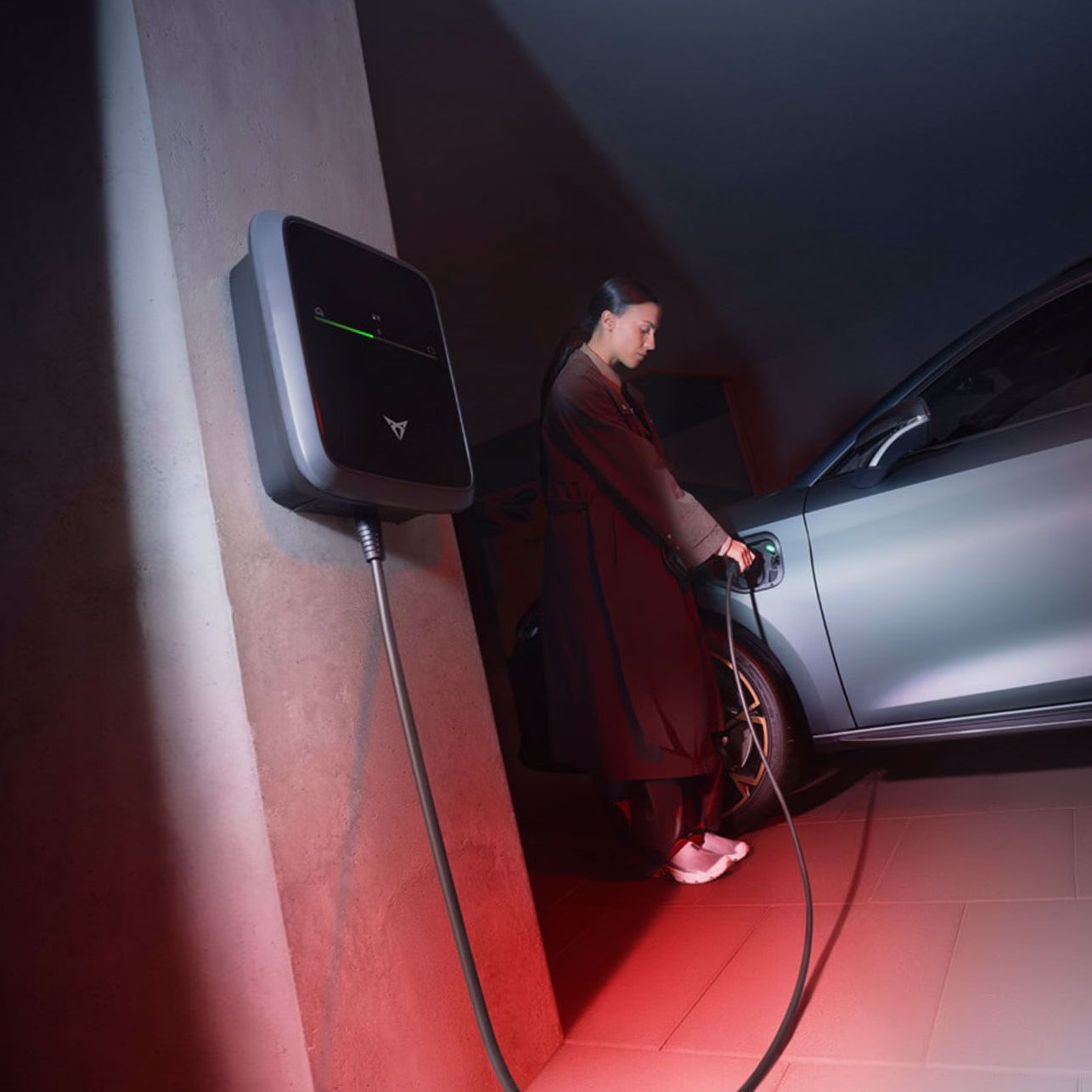 Ženska polni sivi električni avtomobil CUPRA Leon, ki je priključen na polnilno postajo, nameščeno na steni. Okolje je osvetljeno z rdečo in modro svetlobo.