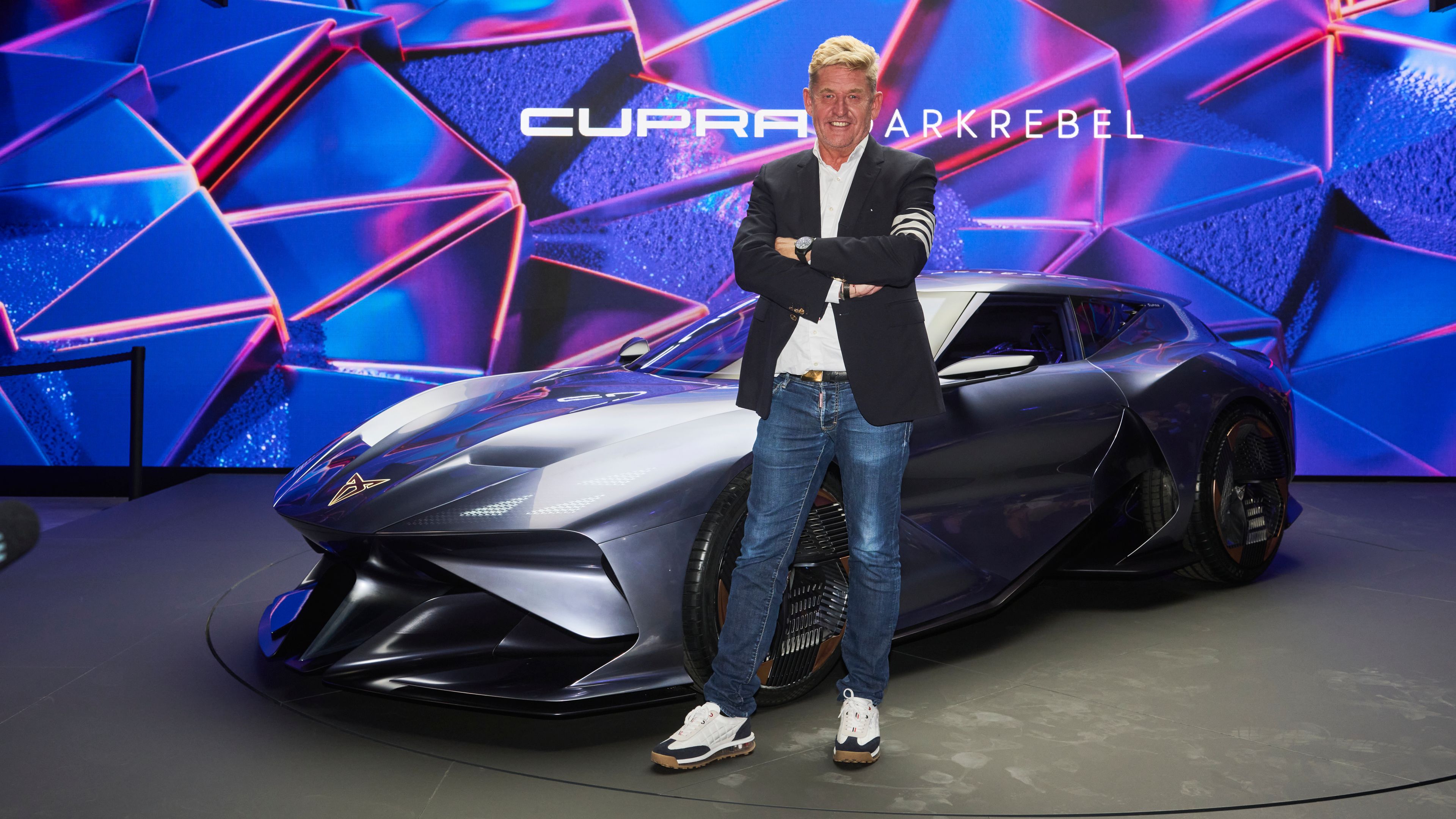 Wayne Griffiths stoji pred sodobnim vozilom CUPRA DarkRebel v salonu. Za avtomobilom je velik zaslon z modro osvetlitvijo.