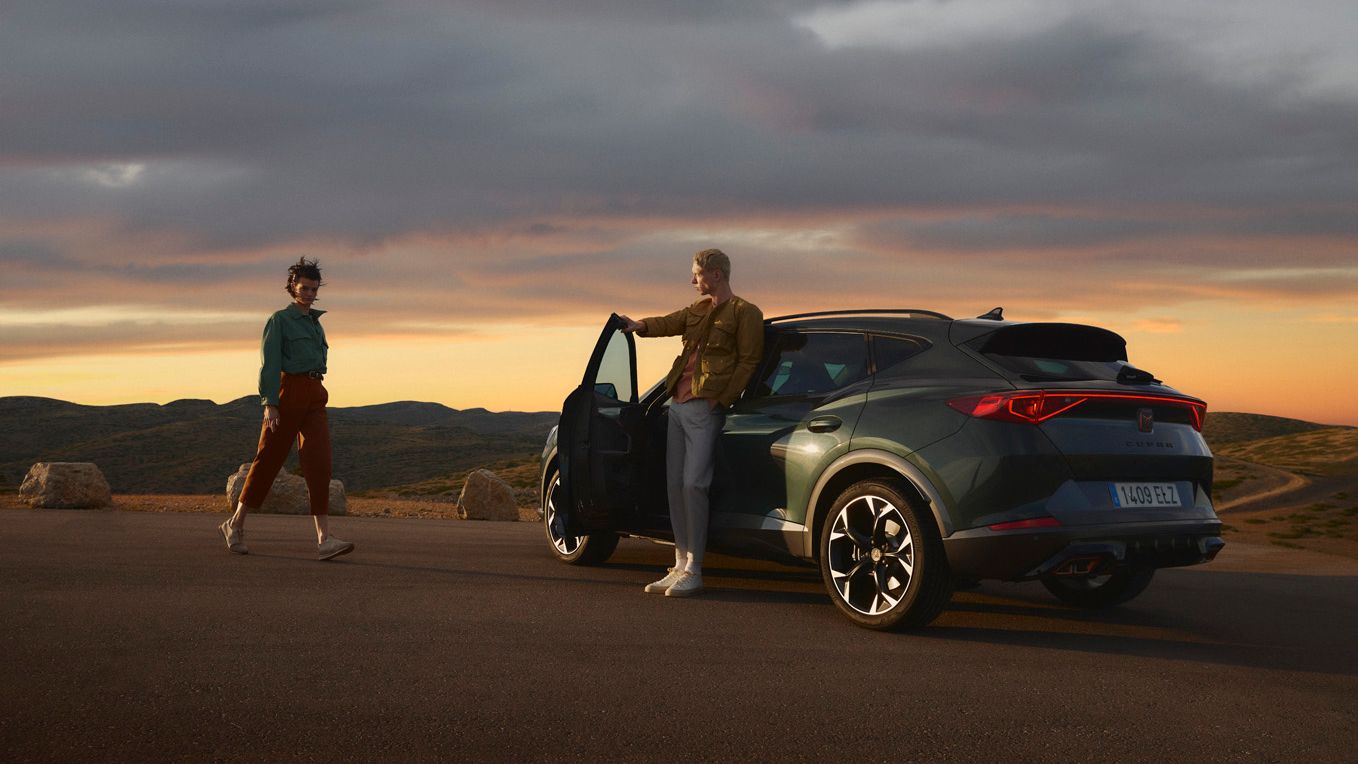 Sivo-zeleni CUPRA Umbrella - Formentor je parkiran ob cesti ob sončnem zahodu v hriboviti pokrajini. Ena oseba stoji ob vozilu z odprtimi vrati, medtem ko druga oseba hodi proti vozilu. 