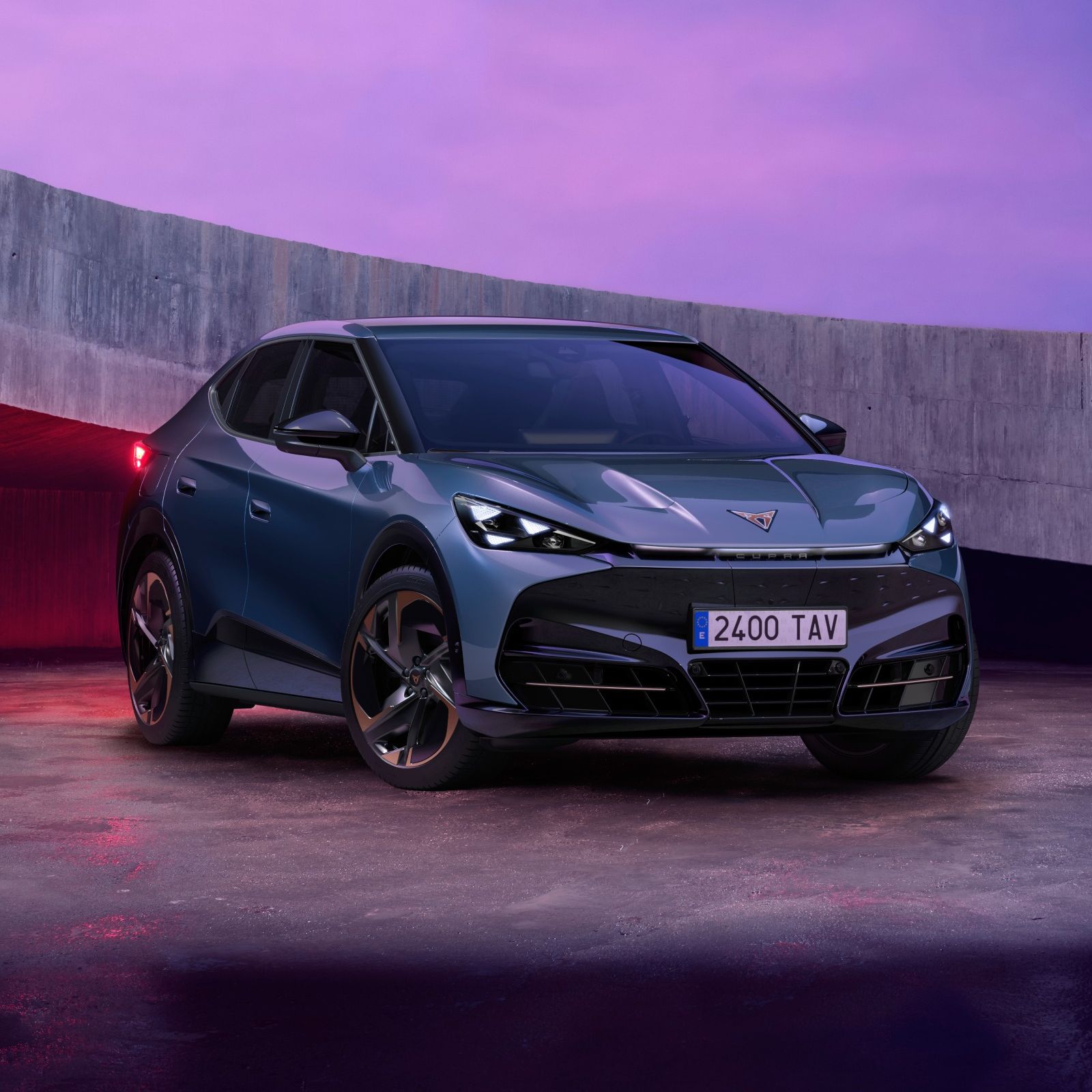 Električni CUPRA TAVASCAN VZ modernega videza, parkiran na betonski površini ob vijoličnemu nebu v ozadju. Avtomobil je modre barve s črnimi detajli. Okolica je osvetljena z mehko svetlobo, ki poudarja linije vozila.